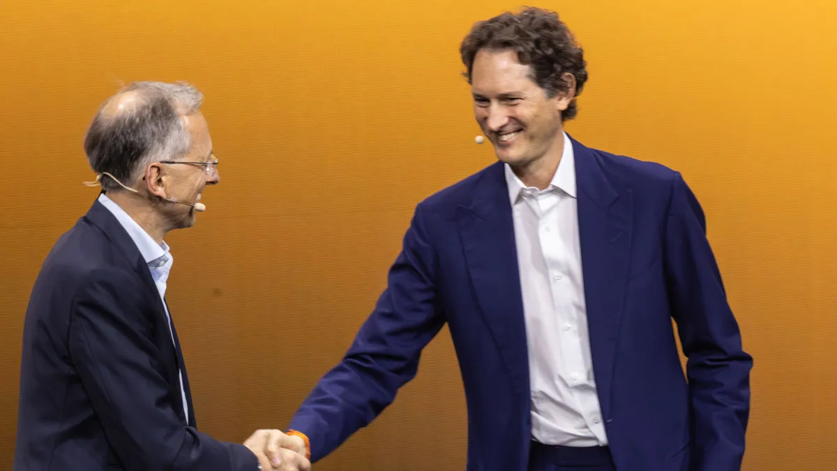 Two men in suits shake hands on a stage with an orange background, microphones on their faces. Wait, that's not necessary.