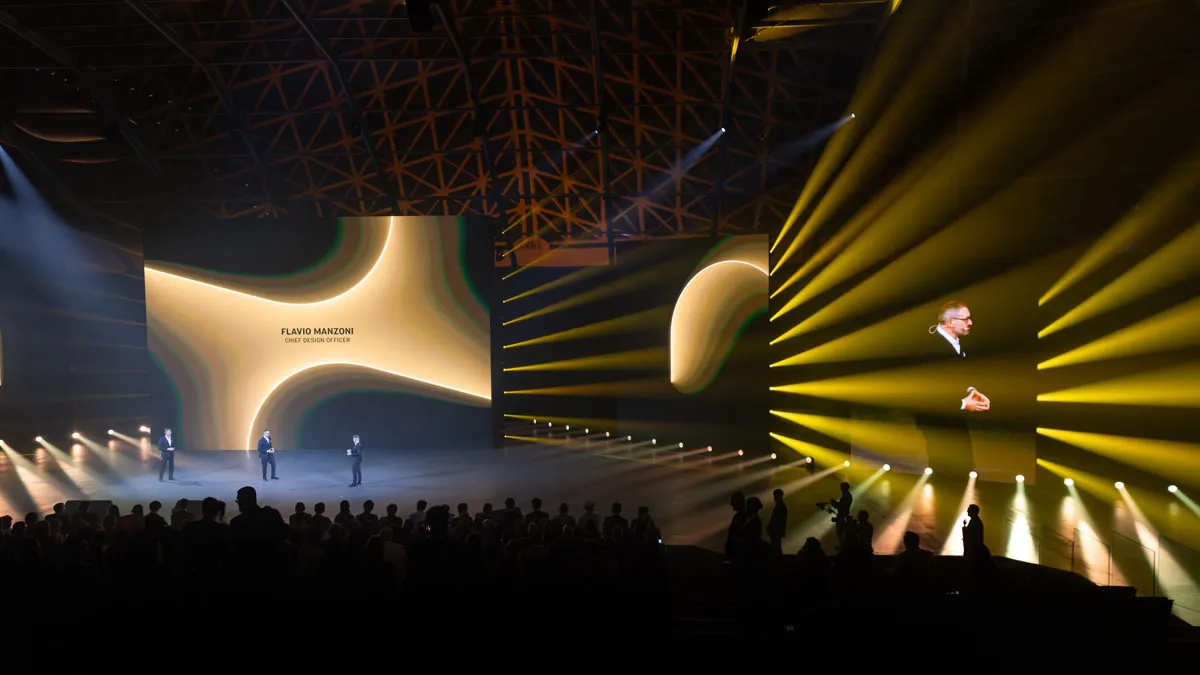 Stage at a conference with three speakers, large abstract screens, and yellow spotlight beams across the audience.