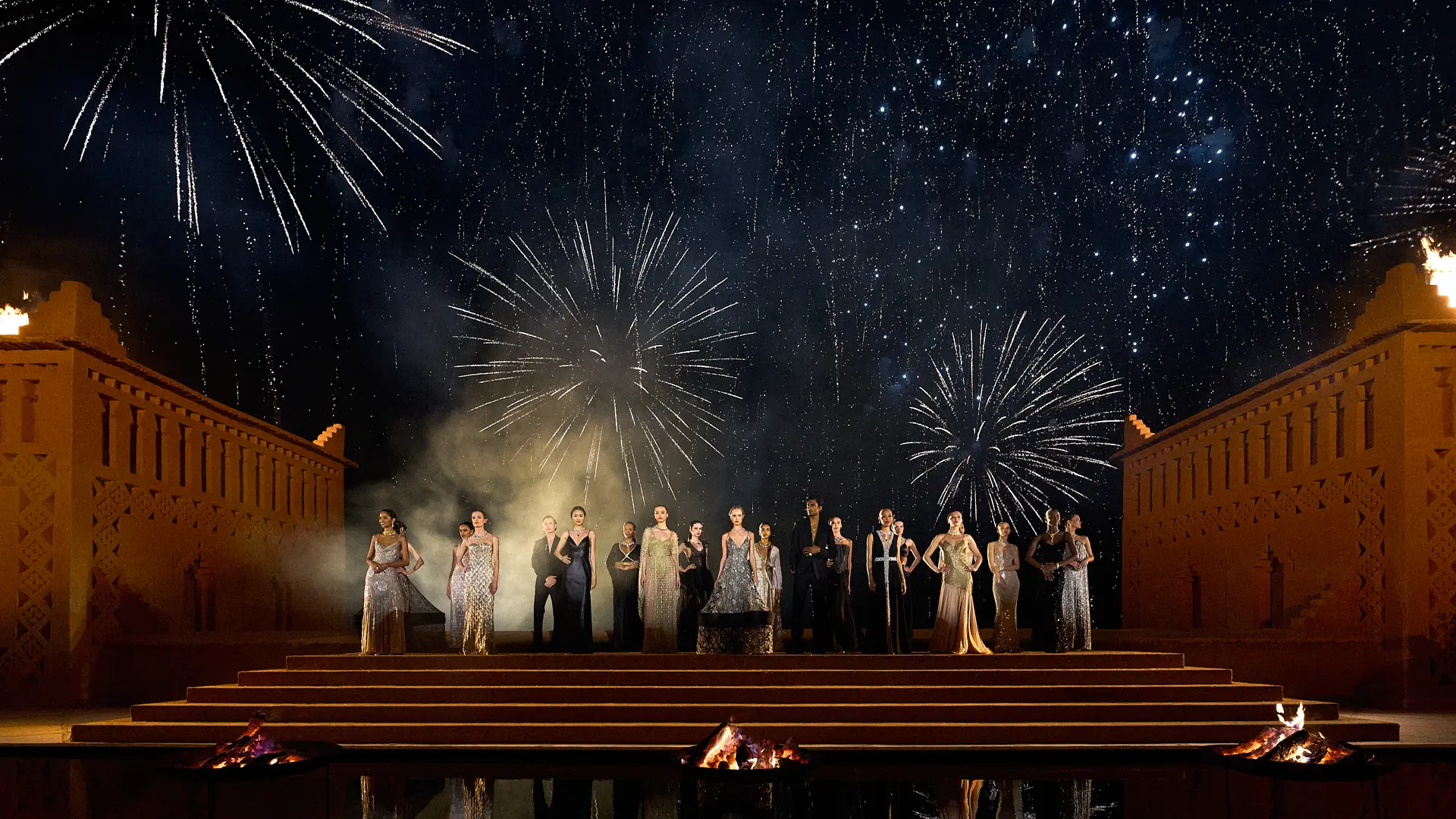 Group of people in formal wear standing on a tiered outdoor stage as fireworks light the night sky above them.