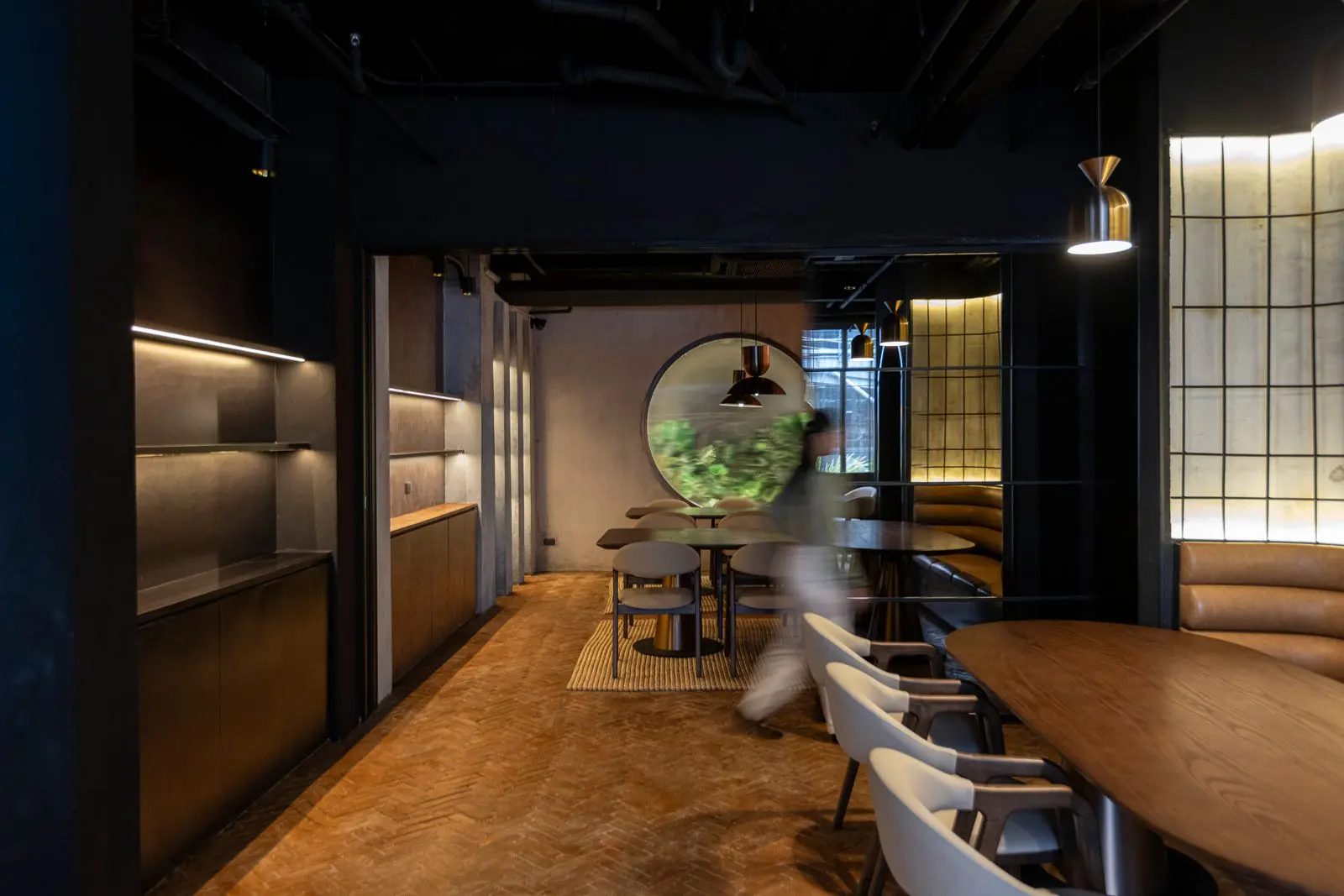 Dark, modern dining room with wooden tables and light-colored chairs, a circular window in the back, and a blurred passerby walking by.