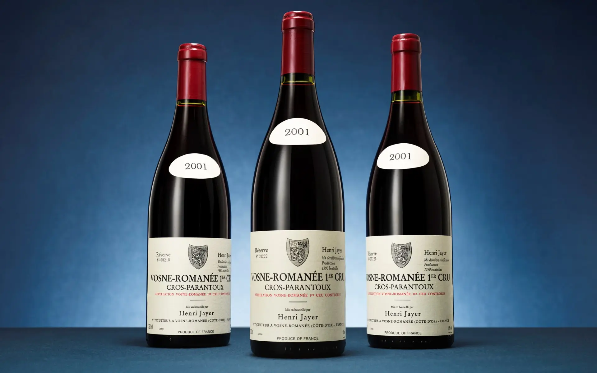 Three wine bottles with red foil caps stand upright on a blue backdrop; each has a white oval sticker reading '2001' above labels that say Vosne-Romanée 1er Cru (wine).