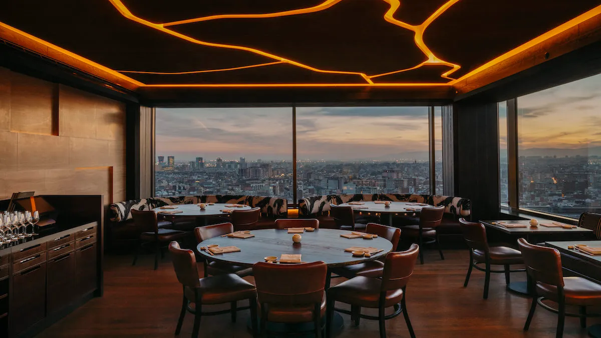 Upscale dining room with round tables, brown leather chairs, and warm orange ceiling lighting overlooking a city skyline at sunset through floor-to-ceiling windows.