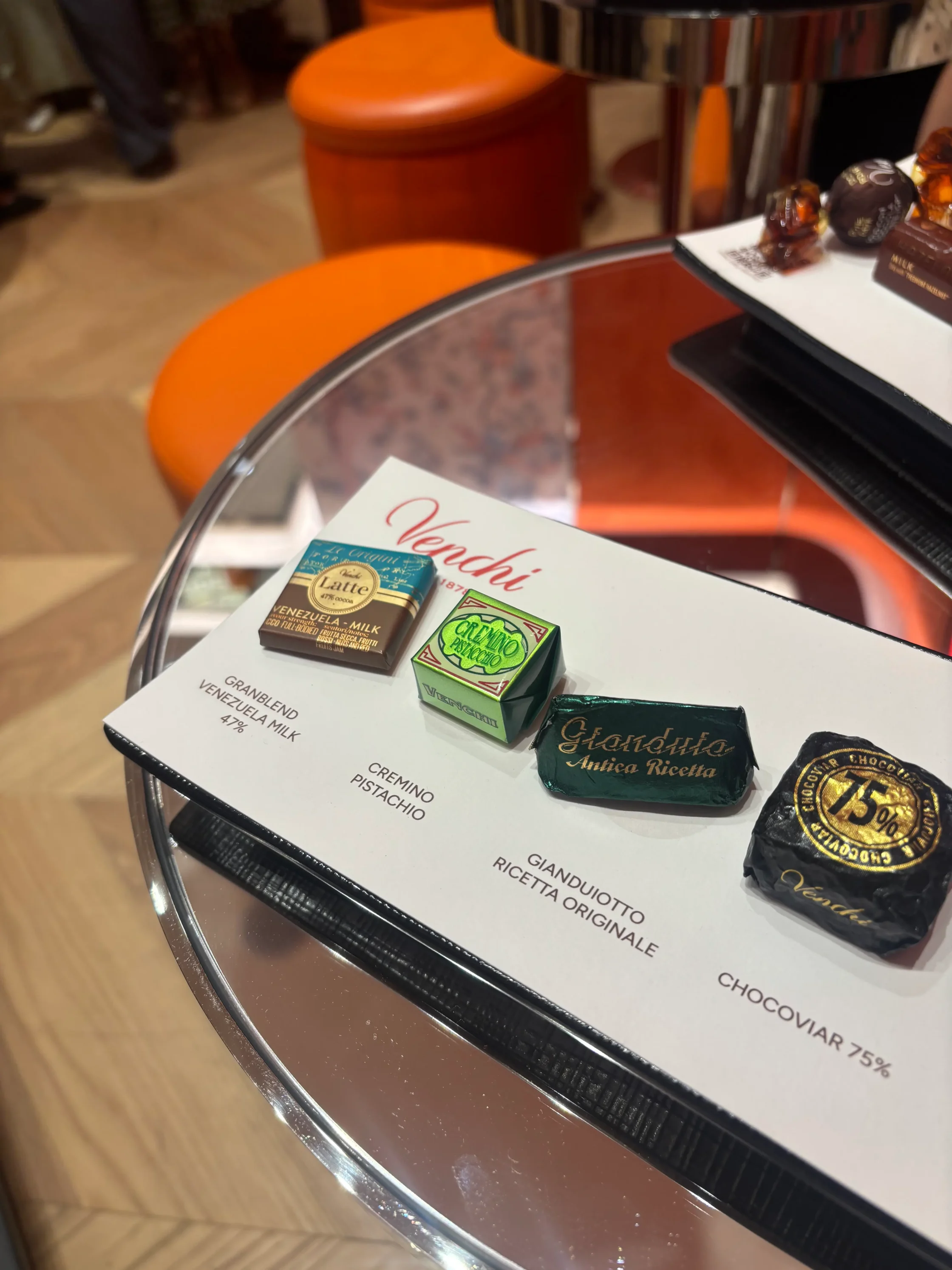 Assorted small chocolate bars placed on a white menu card inside a curved glass display, with Venchi and Gianduotto labels visible.