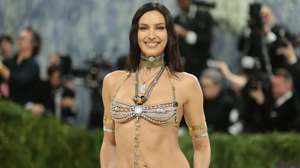 Woman on a red carpet wearing a jeweled, harness-style top and layered necklaces, with photographers in the background.
