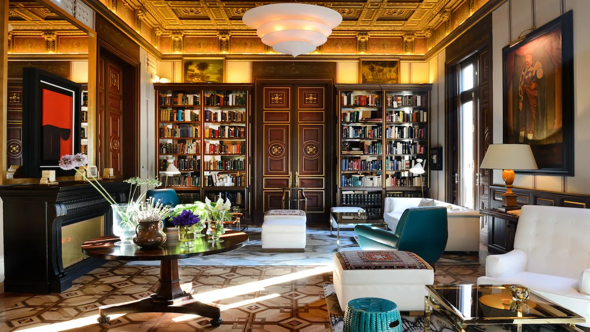 Luxurious library lounge with ornate gold-trimmed ceiling, tall bookshelves, and a central wooden door behind seating area.