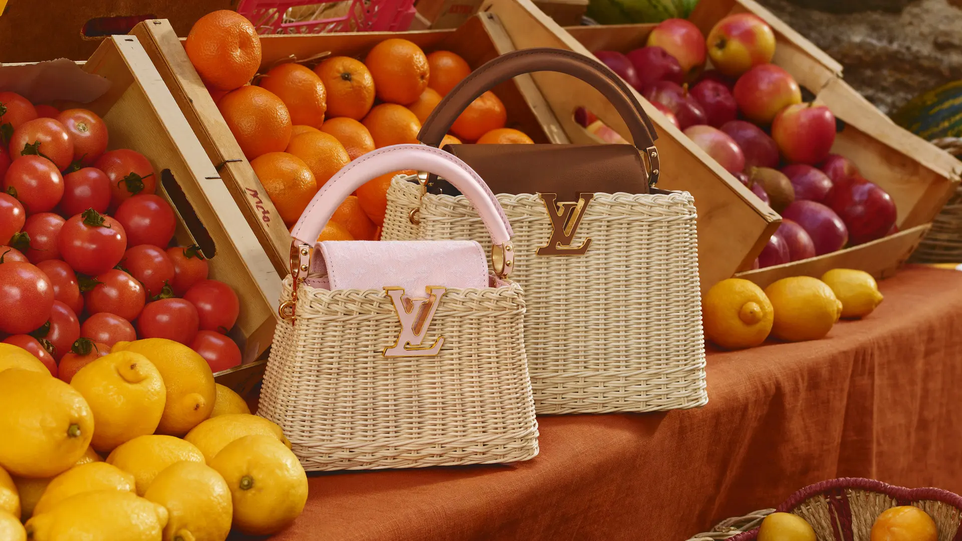 Three Louis Vuitton wicker handbags displayed on a market fruit stand among oranges, lemons, and apples.