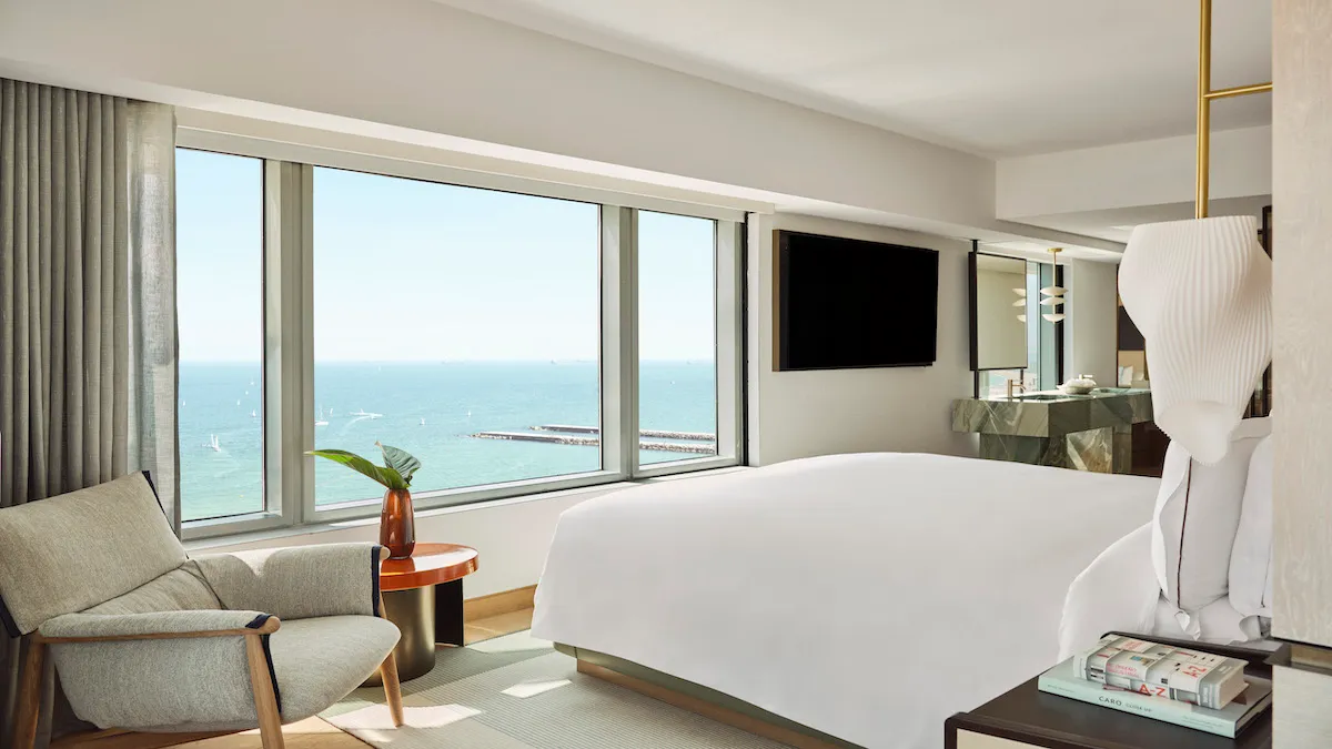 Hotel room with a large window showing an ocean view, a white bed, and a sitting area by the window.