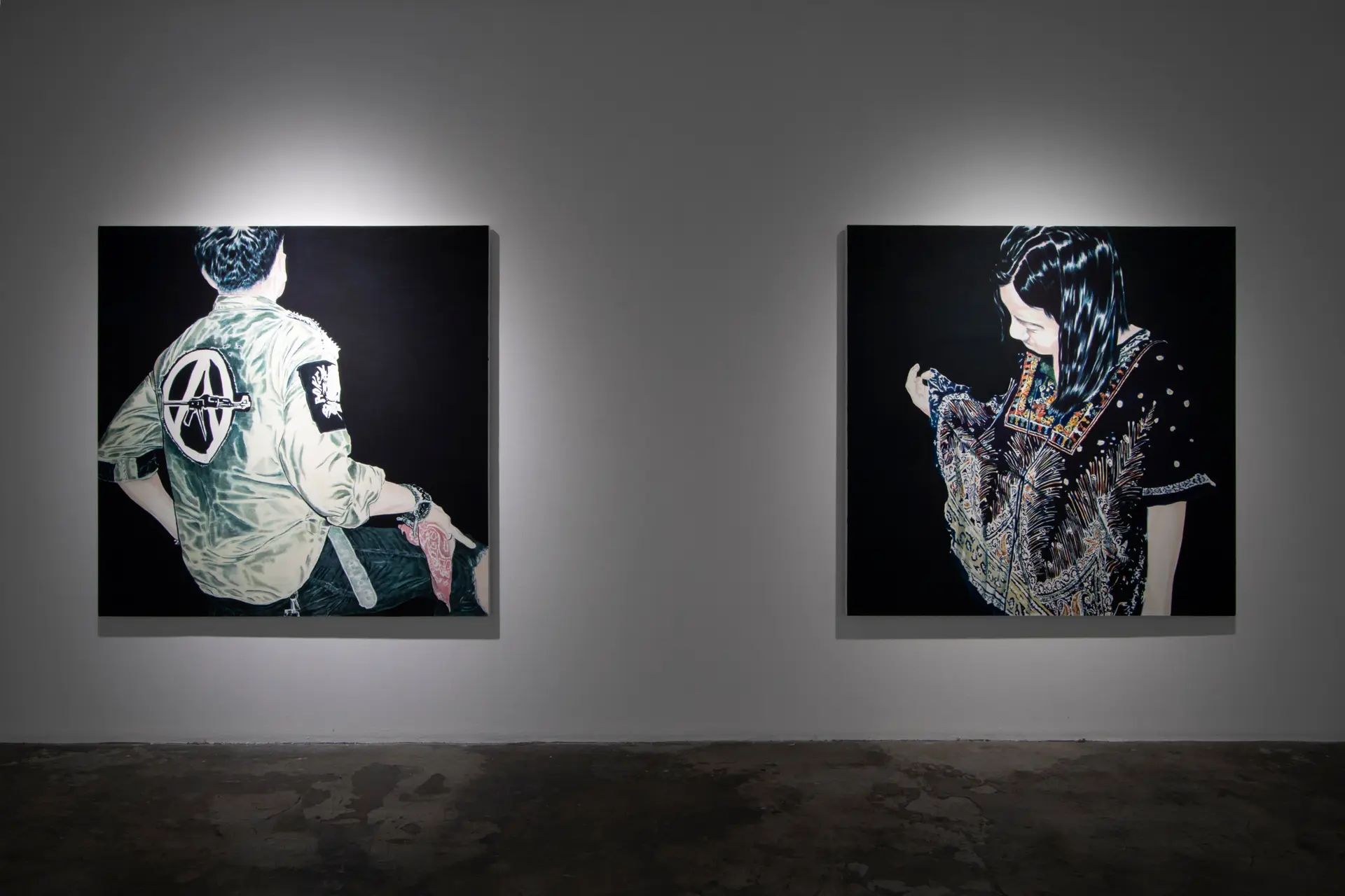 Two large framed portraits on a dim gallery wall under spotlights: left shows a person viewed from behind in a light jacket with patches; right shows a woman in a patterned dress looking down.