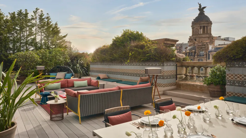 Rooftop terrace with colorful cushioned sofas, a long dining table set for a meal, and potted plants, with a historic domed building in the background.