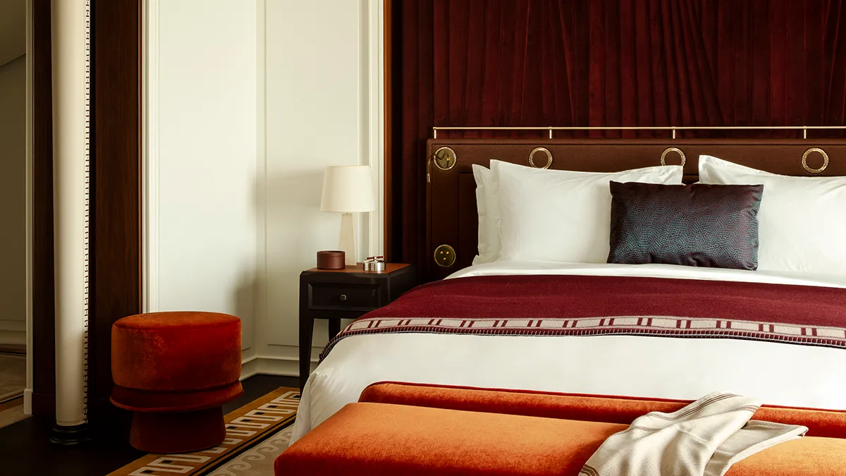 Luxurious hotel bedroom with a king bed, white linens, burgundy headboard, and an orange ottoman.