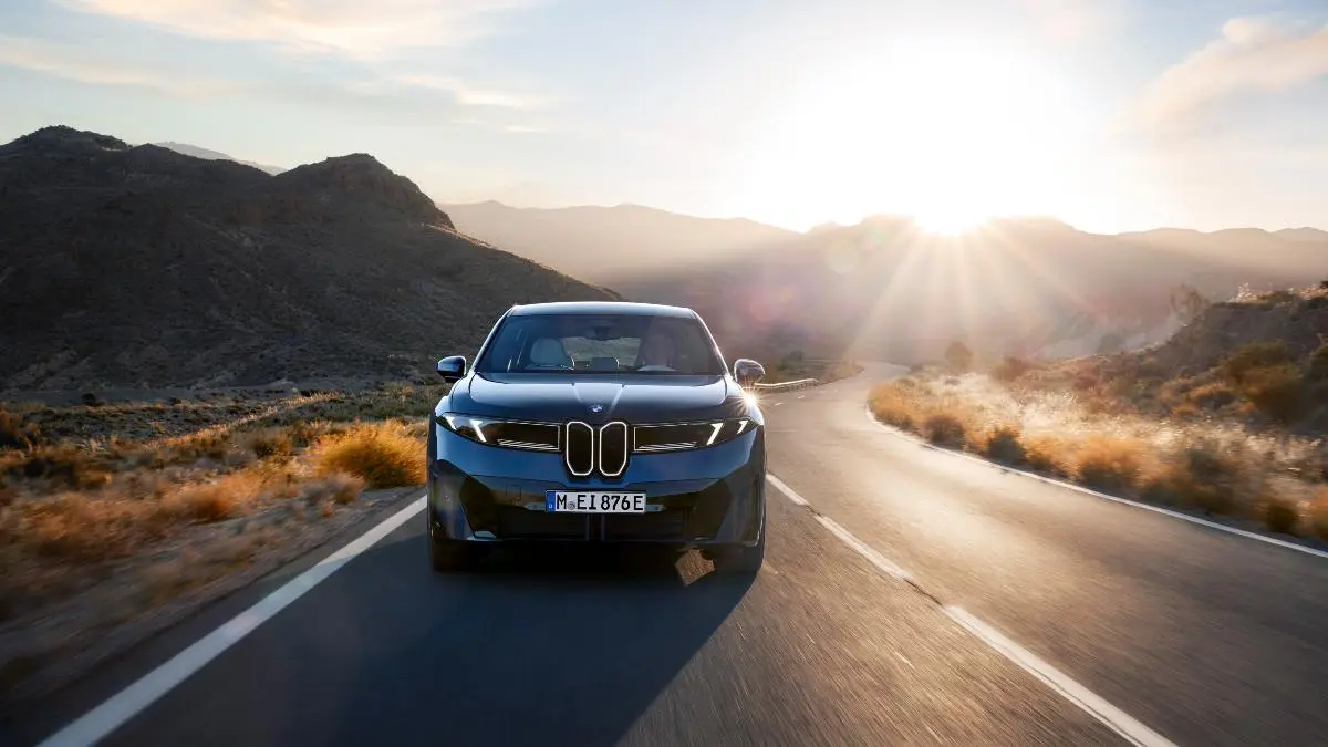 Front view of a dark BMW driving on a winding desert road at sunset with rocky hills in the background.