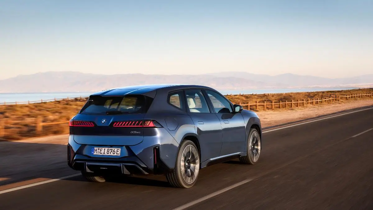 Blue BMW electric SUV seen from the rear three-quarter on a coastal highway with a sea and mountains in the distance, sunset lighting.