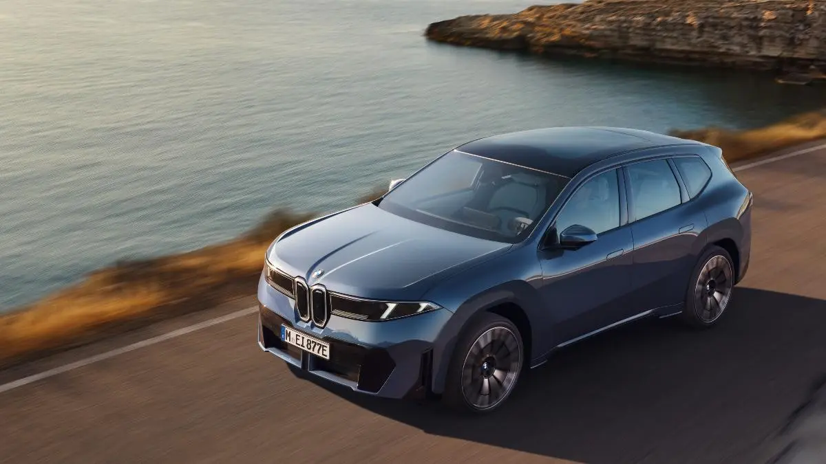 Front three-quarter view of a blue BMW SUV driving along a coastal highway with the sea on the left and rocky cliffs in the background.