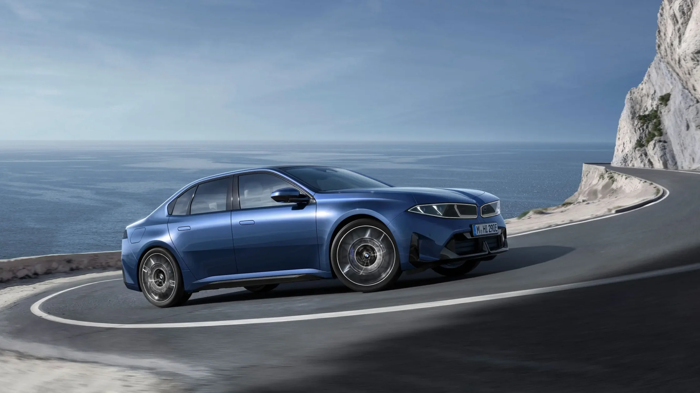Blue BMW i3 driving along a coastal mountain road with a sea view and rocky cliff on the right.