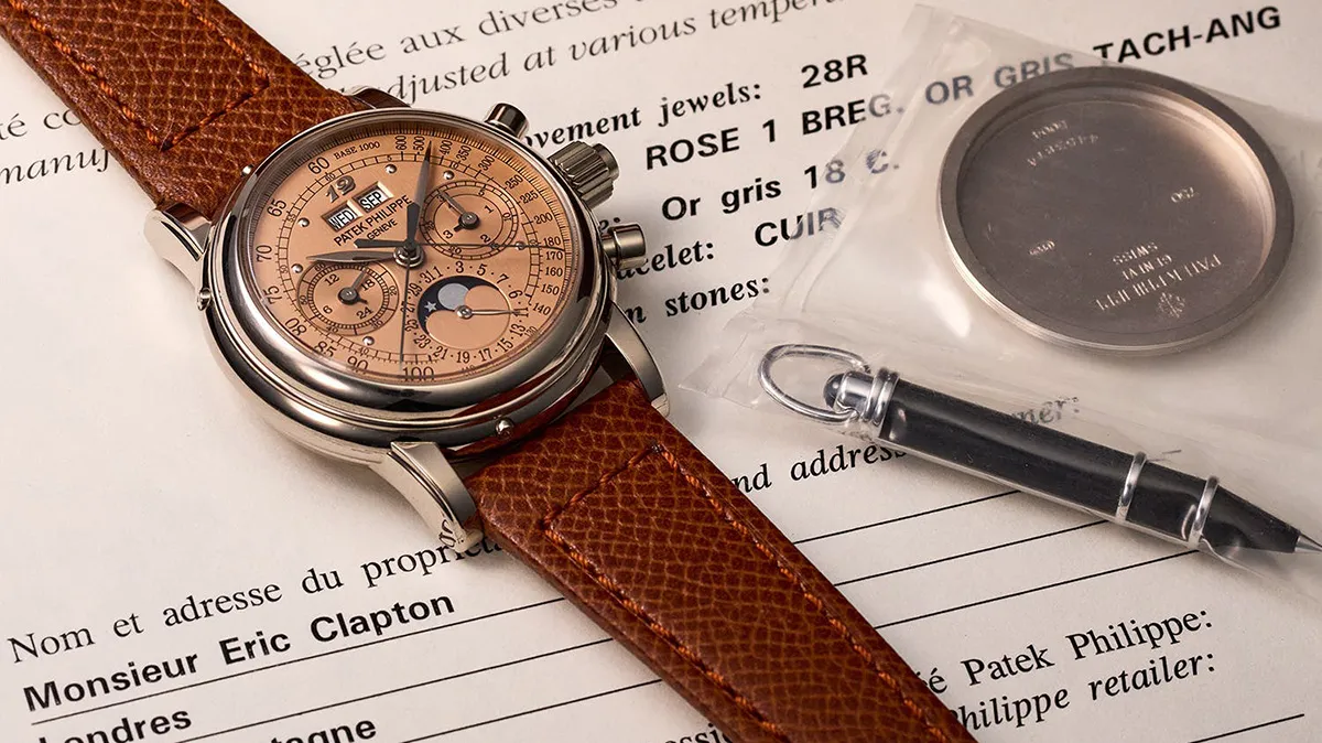 Patek Philippe chronograph watch with a brown leather strap resting on a French document, with a fountain pen and a metal case nearby.