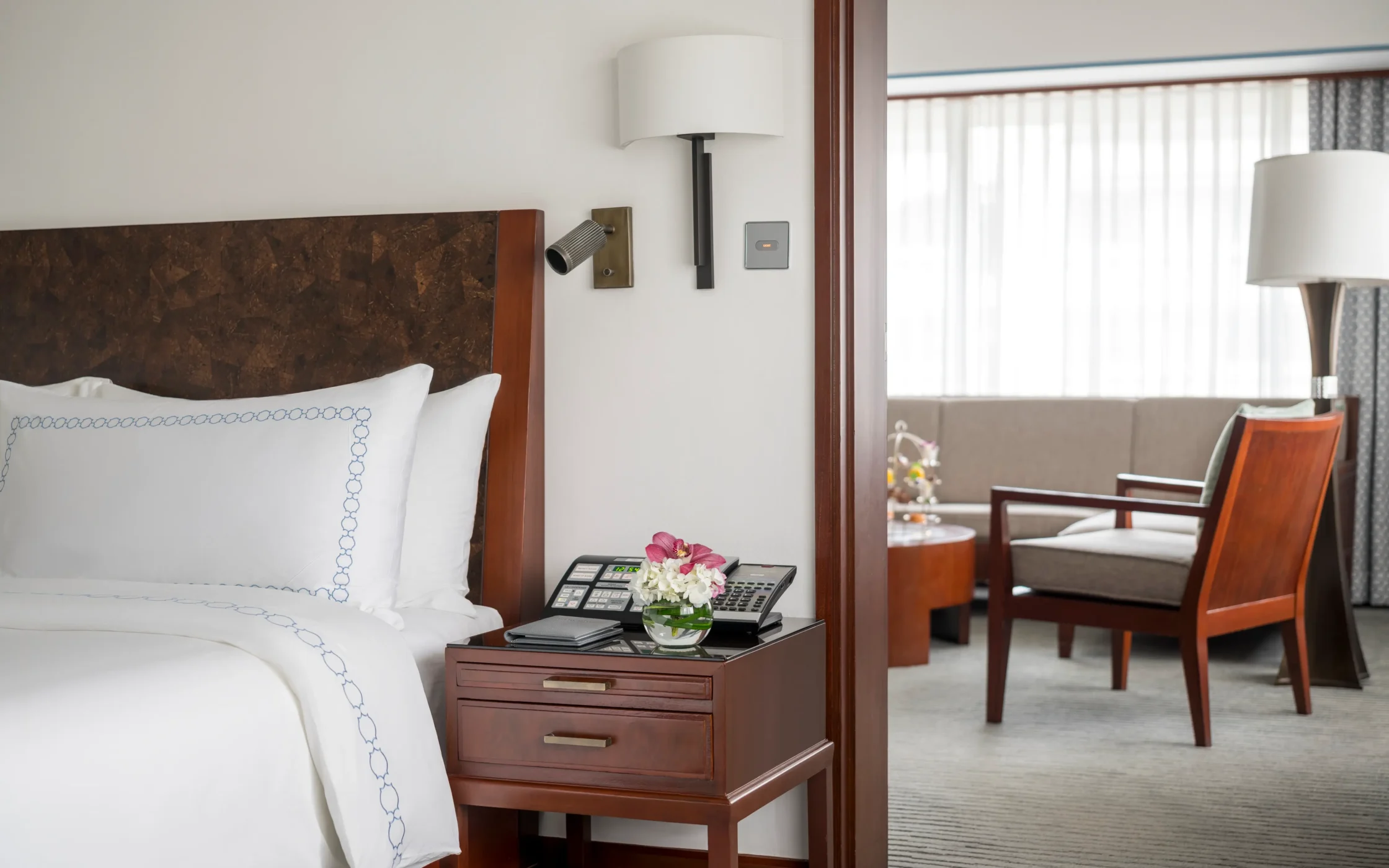 Hotel room with a white bed and wooden headboard, a nightstand holding a phone and flowers, and a doorway to a seating area in the background.