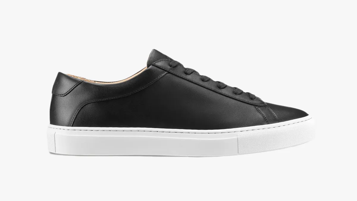 Black low-top sneaker with a white thick sole and black laces on a plain background, side view.