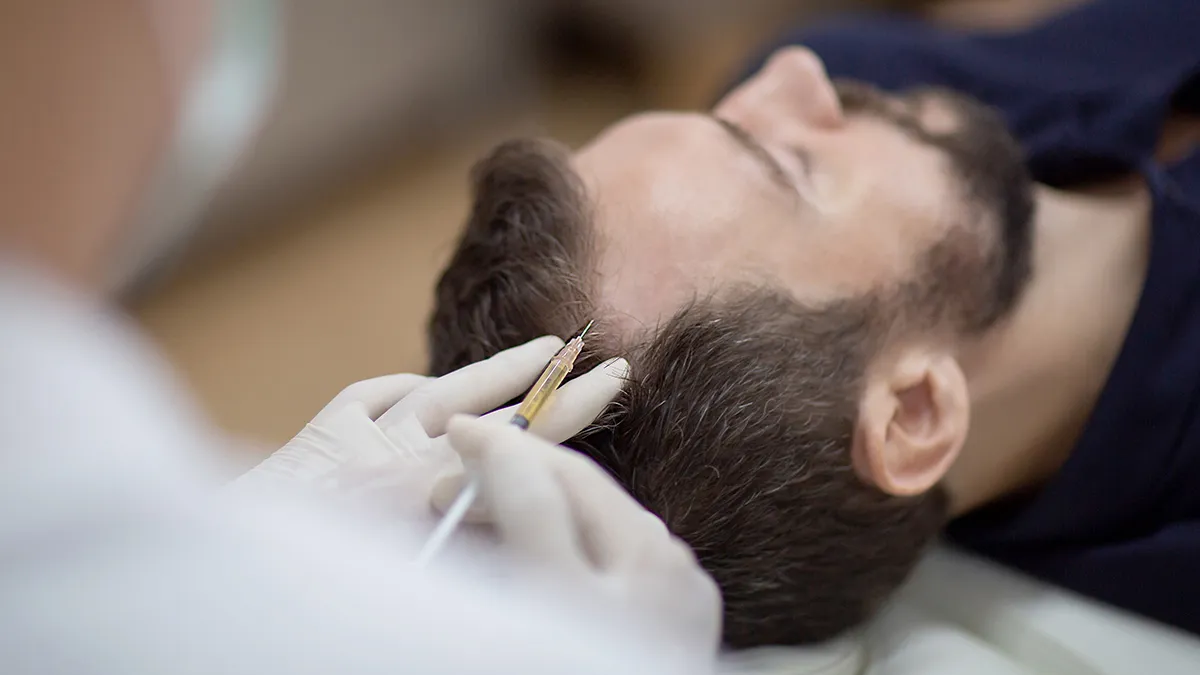 Healthcare worker administers an injection to a man lying on his back, near the neck/jaw area, with gloved hands and a syringe.