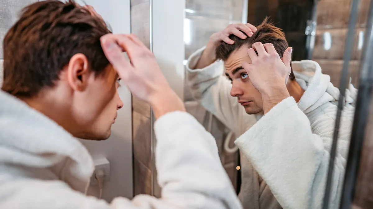 Man in a white bathrobe looks at himself in the bathroom mirror, styling his hair with both hands