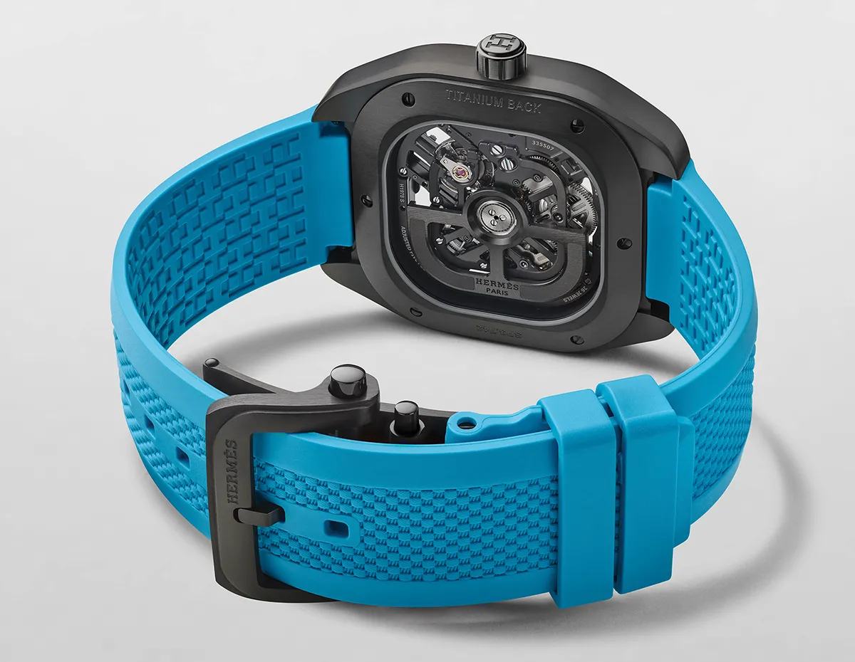 Back view of a black-toned wristwatch with a bright blue rubber strap and visible mechanical movement through the case back, Hermes Paris branding.