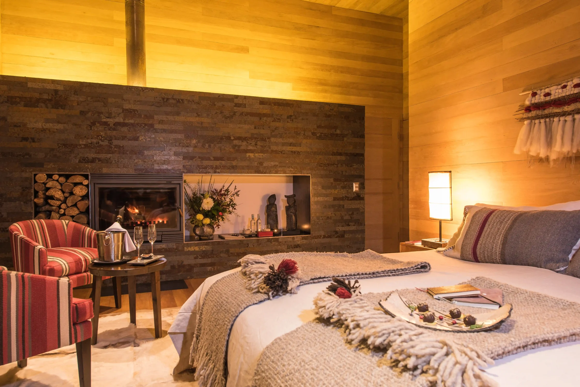 Cozy hotel room with a lit stone fireplace, wood panel walls, and a striped red chair beside a small table with a champagne bucket and glasses.
