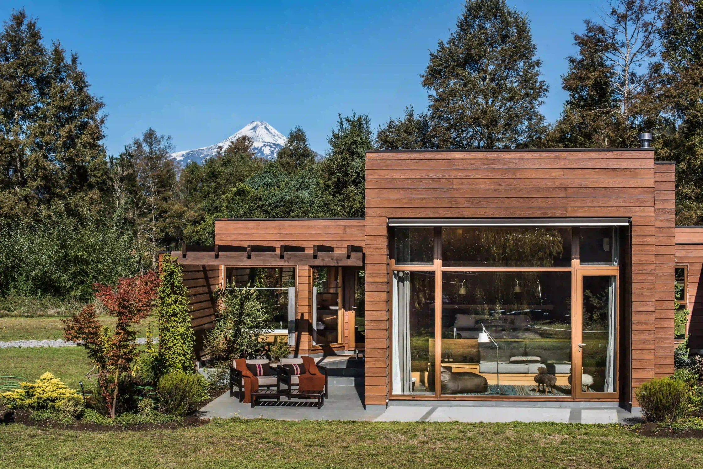 Modern wooden house with large glass doors and a seating area, set in a landscaped yard with a snow-capped mountain in the background.