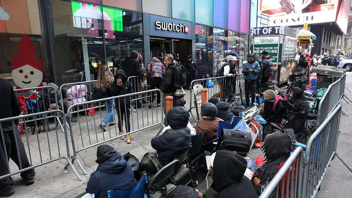 People wait in a long queue behind metal barricades outside a colorful storefront on a busy city street with shoppers nearby.