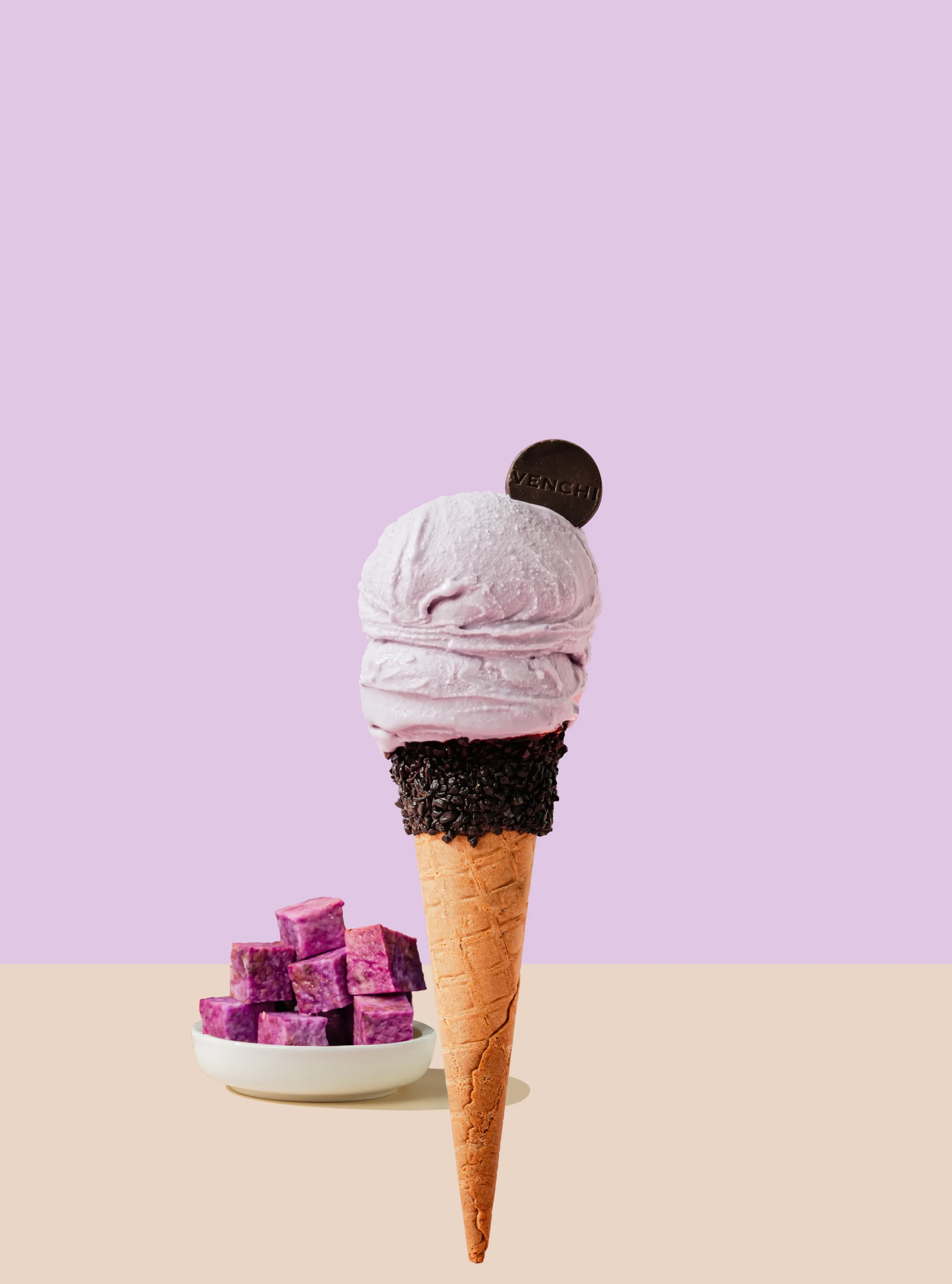 Ice cream cone with a pink scoop and chocolate-dipped base, paired with a bowl of purple cubes on a lavender background.