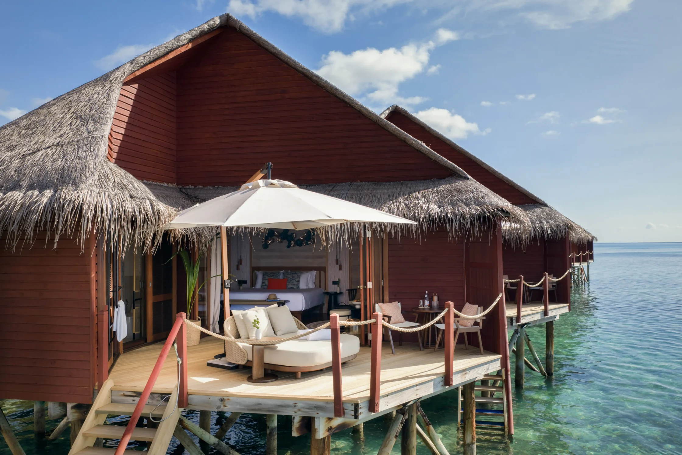 Overwater bungalow with thatched roof and red walls, deck seating, and an umbrella over turquoise water.