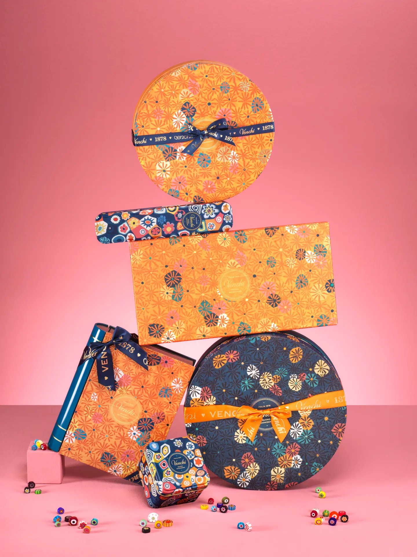Brightly patterned gift boxes and ribbons stacked on a pink background into a festive display.