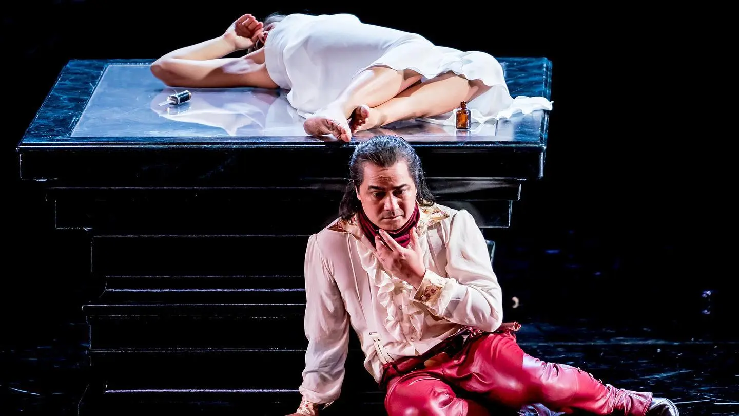 Theatrical scene: a man in a white shirt and red pants sits on the floor beside a raised platform with a body under a white sheet; a bottle and mic lie on the platform.