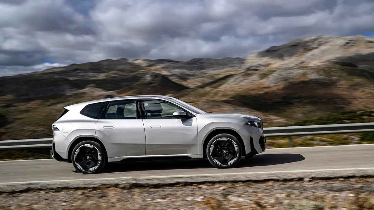 Silver SUV driving along a mountain road with a guardrail and rocky hills under cloudy skies.