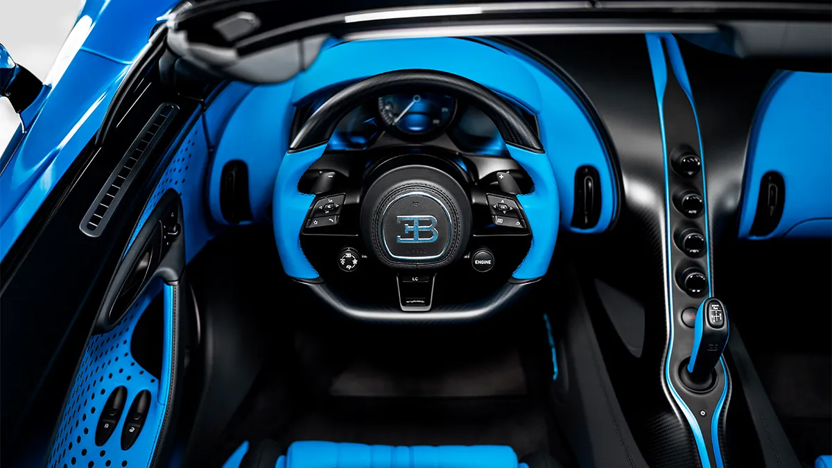 Blue leather interior of a luxury sports car with a circular steering wheel and carbon-fiber accents featuring a Bugatti emblem at the center.