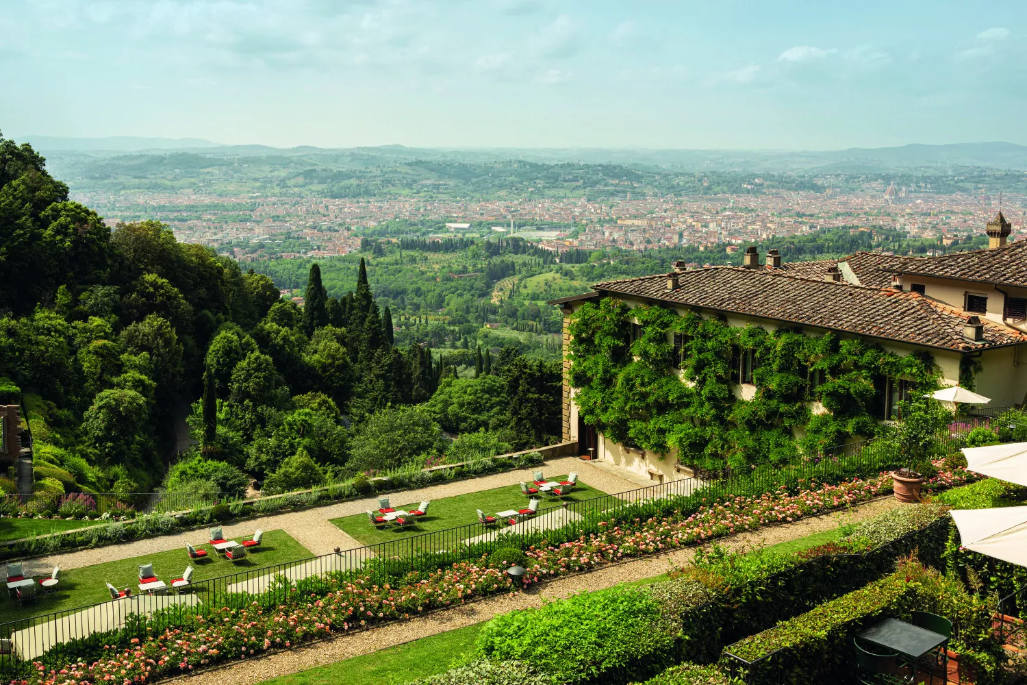 Villa with ivy-covered wall and a manicured garden overlooking a valley and distant city skyline, with red chaise lounges on the lawn.