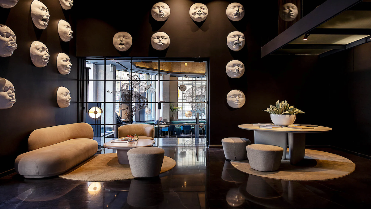Modern lounge with black walls covered in white moon-face sculptures, beige curved sofa, round coffee table, and gray ottomans on circular rug.