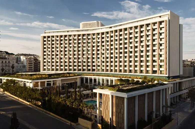 Large curved modern hotel building with many balconies and a rooftop garden visible at the top, viewed from the street level outside.