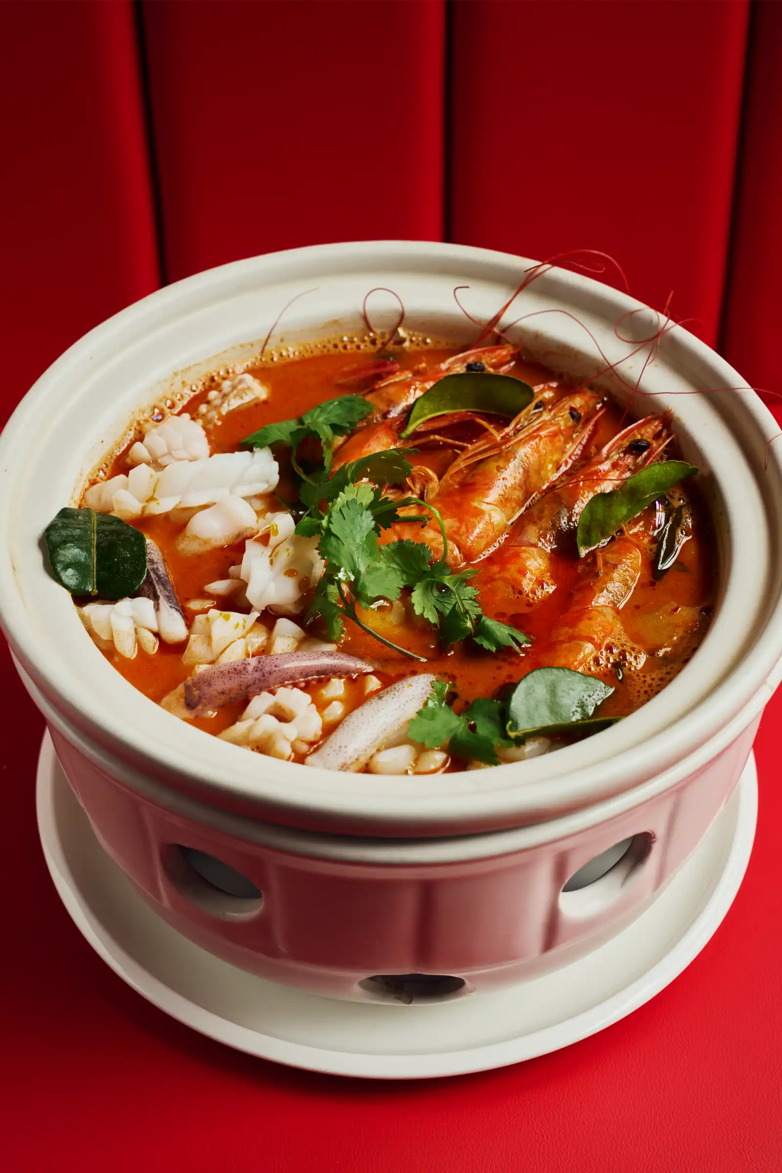 Seafood hot pot with shrimp, squid, and cilantro in a red spicy broth, served in a white ceramic pot on a red surface.