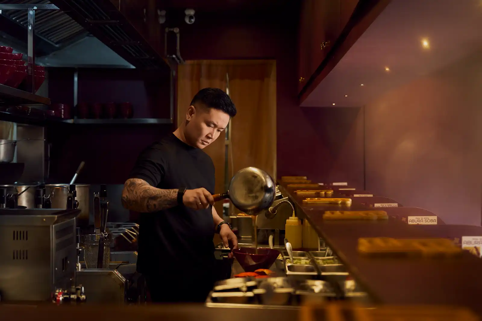 Chef in a dark kitchen plating food, pouring from a pan over a bowl at a ramen counter.