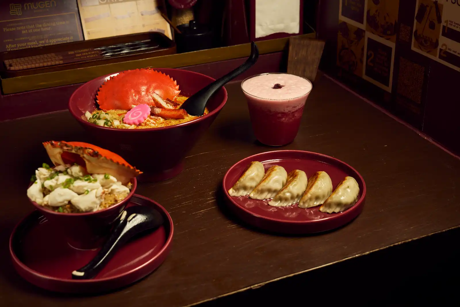 Ramen meal with crab leg, pink swirl fish cake, and toppings in a red bowl, plus a pink frothy drink and a plate of dumplings on a dark table.