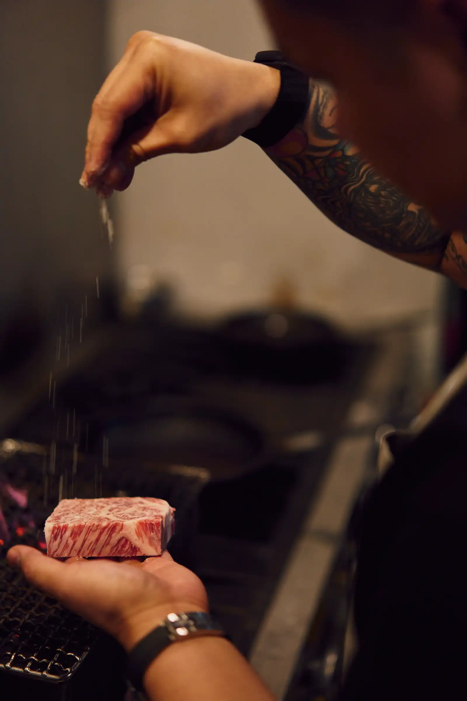 Close-up of a tattooed forearm holding a marbled raw steak cube over a grill.