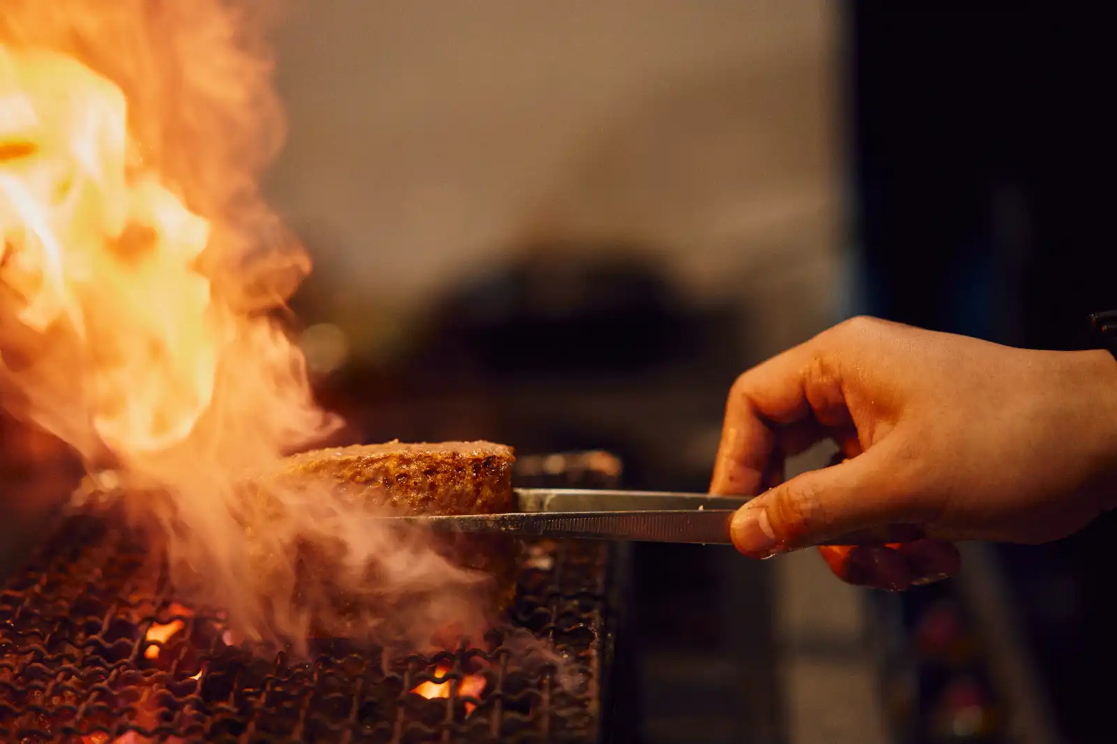 Person using tongs to grill a hamburger patty over open flames on a charcoal grill