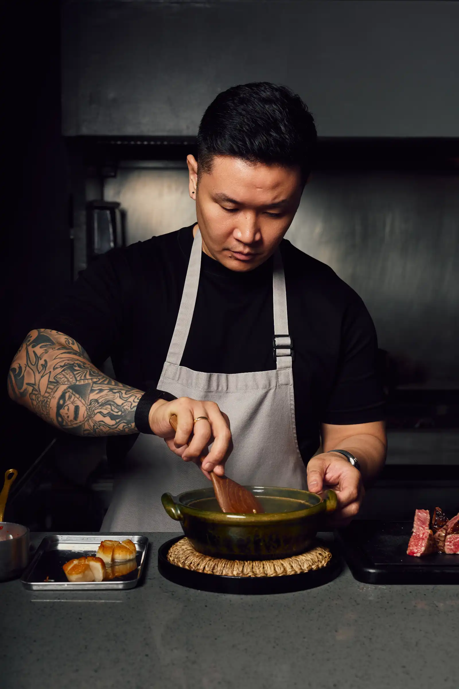 Male chef with a tattooed forearm, wearing a grey apron, stirring contents in a green pot on the stove.
