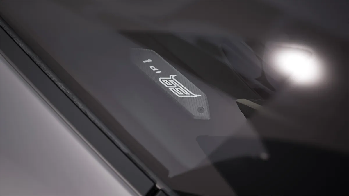 Close-up of a Lamborghini Revuelto NA63 windshield with a small gray badge/sticker on the glass, with a bright headlight blurred in the background.