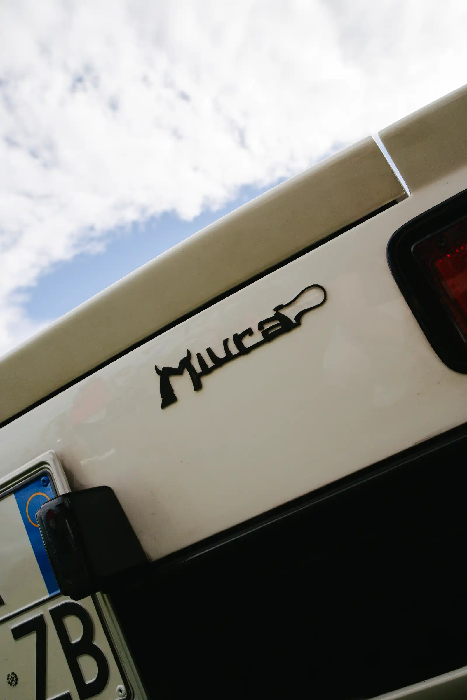 Close-up of a white vehicle rear with a partial black logo and red taillight, against a blue sky with clouds.