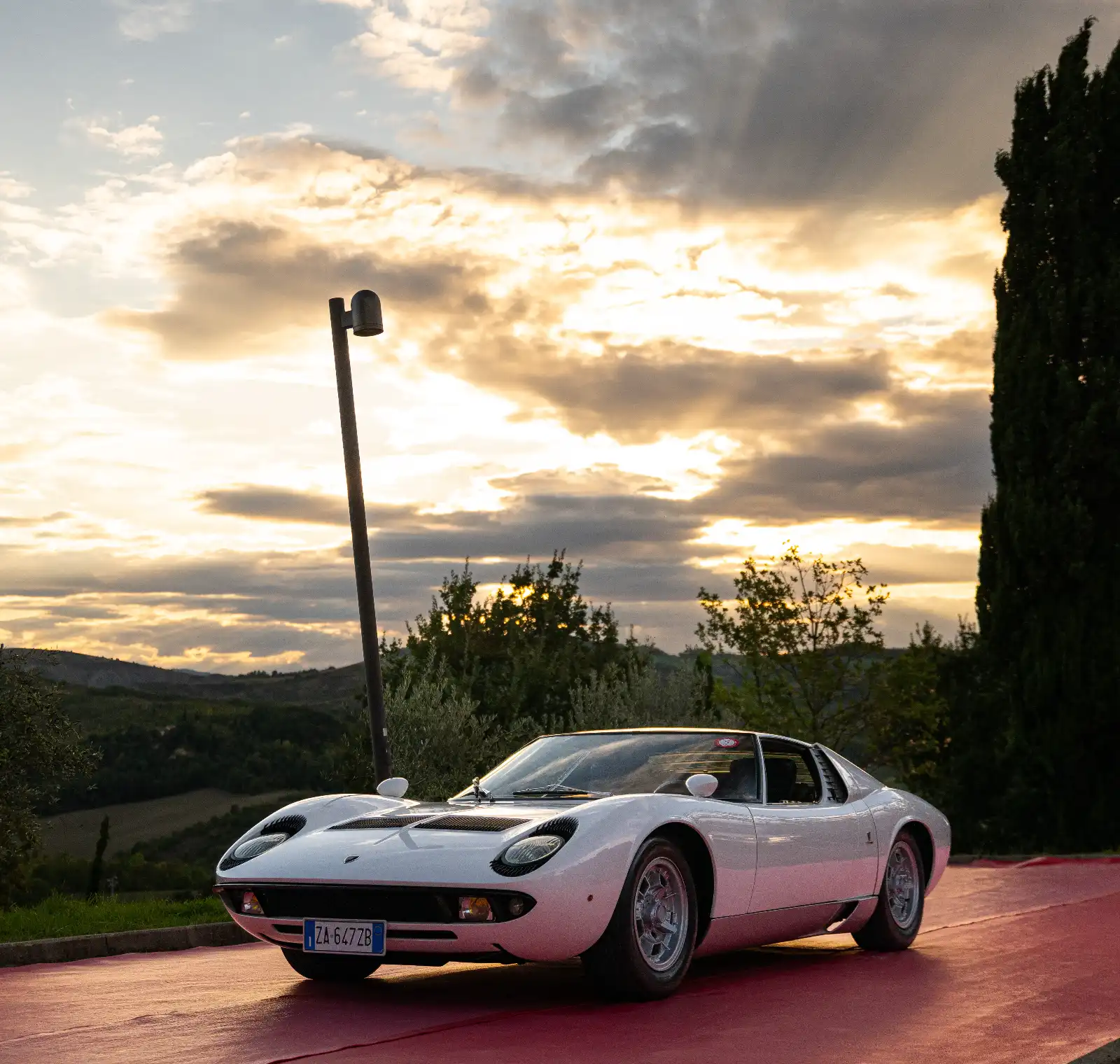 White sports car parked on a red pavement at sunset, leaning street lamp nearby and hills in the distance.