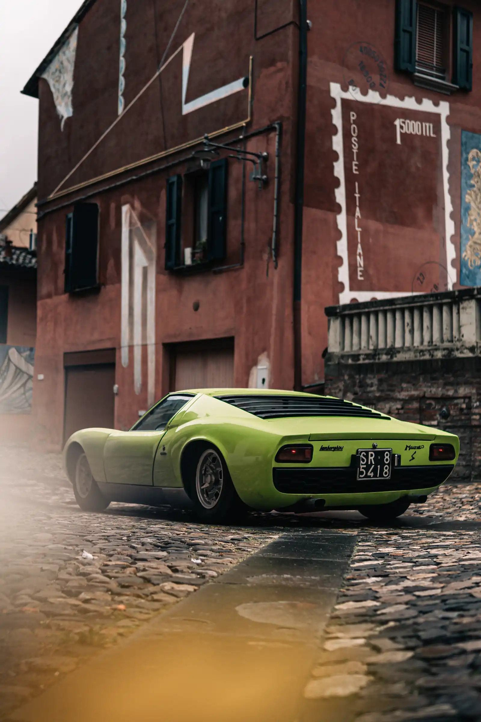 Lime-green Lamborghini parked on a cobblestone street beside a red building with a large stamp-themed mural on the wall.