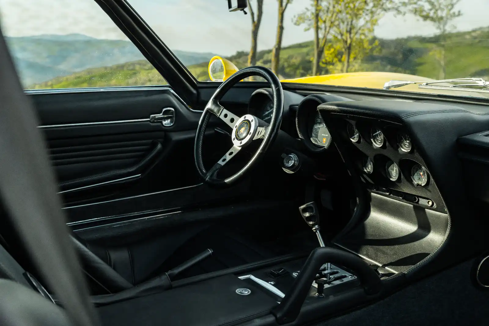 Interior of a classic car with a black dashboard, perforated steering wheel, and yellow exterior visible through the windshield,