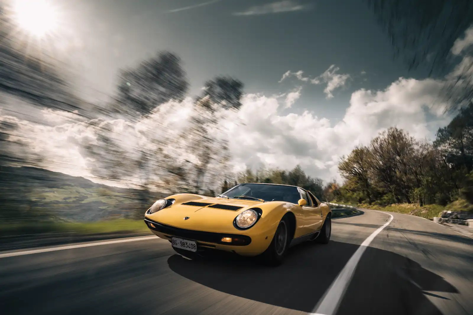 Yellow sports car races along a winding mountain road, motion blur emphasizing speed under a cloudy sky.