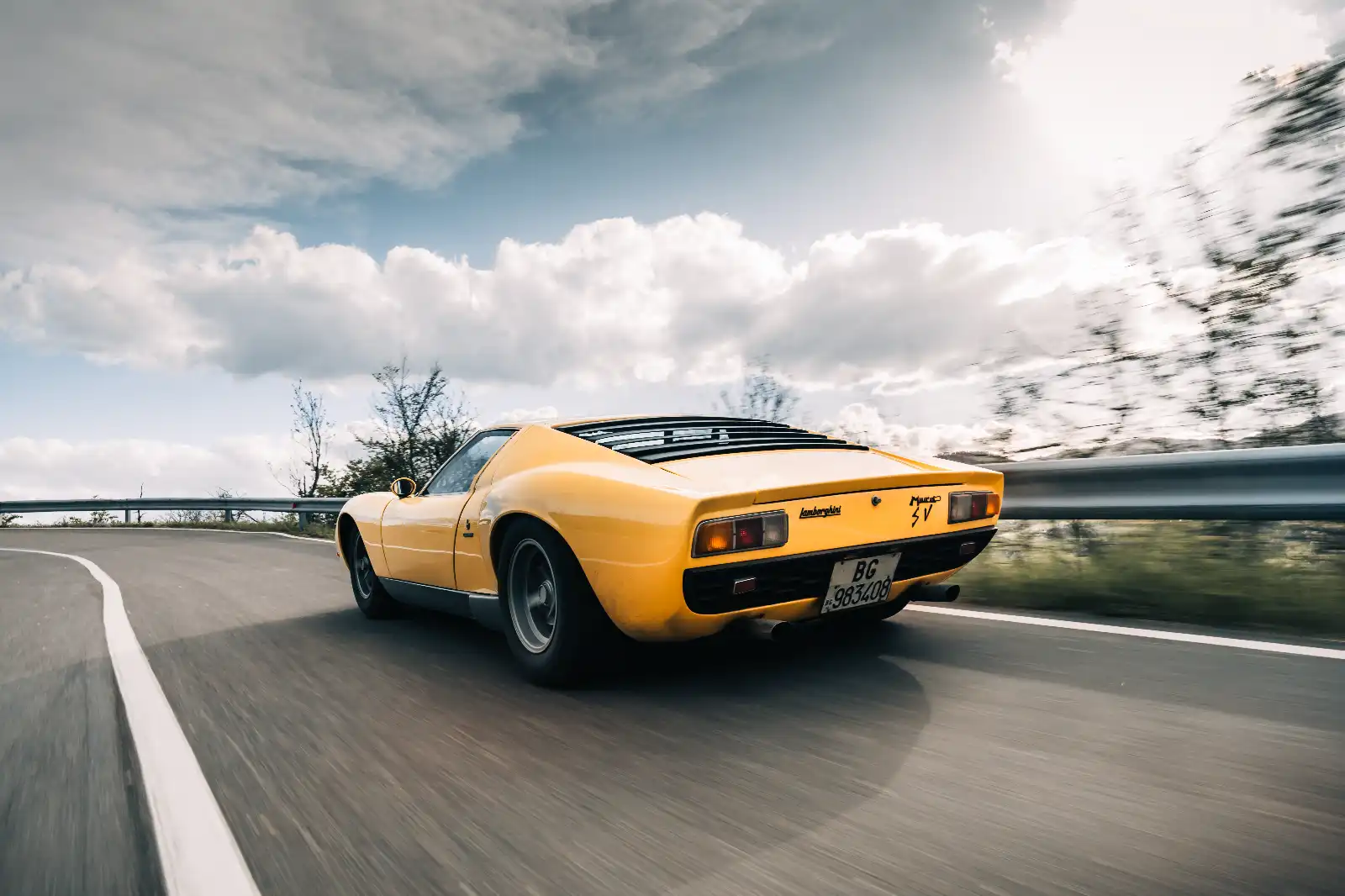 Rear view of a yellow Lamborghini Miura on a winding road with a guardrail and blue sky above I wisps of clouds.”] ,