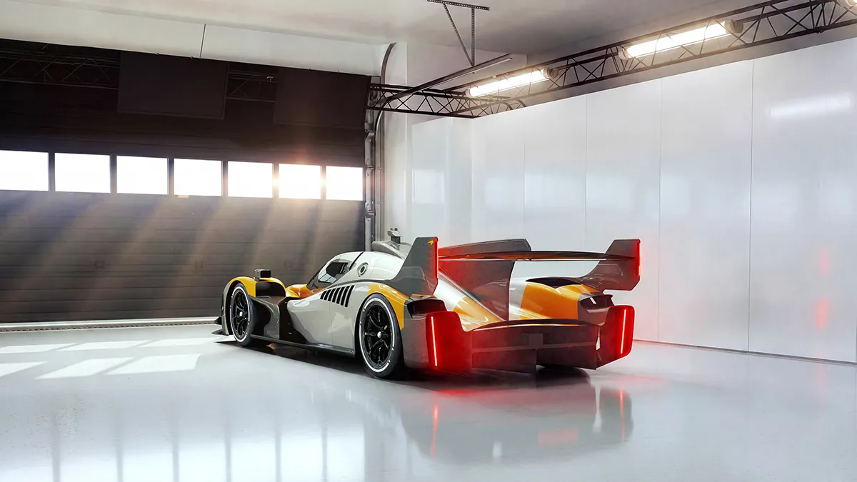 Orange and white prototype race car with a large rear wing parked in a bright indoor garage, sunlight streaming through windows, red taillights glowing.